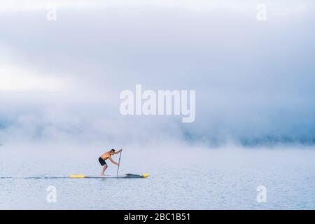 Mann steht auf Paddelsurfen auf einem See im Nebel Stockfoto