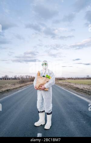 Mann, der Schutzanzug und Maske trägt, hält Lebensmitteltasche auf einer Landstraße Stockfoto