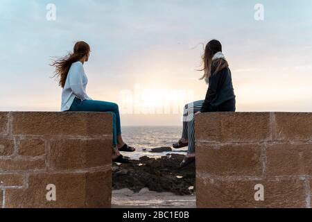 Zwei junge Frauen sitzen an der Wand am Strand und beobachten den Sonnenuntergang in Essaouira, Marokko Stockfoto