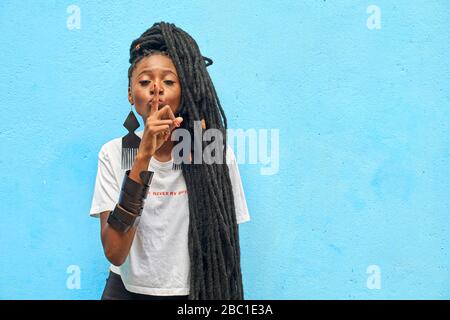 Porträt einer Frau mit langen Dreadlocks, die das Schweigeminde mit dem Finger vor der türkisfarbenen Wand machen Stockfoto