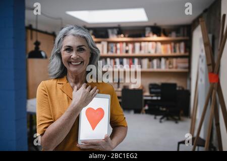 Lächelnde grauhaarige Frau, die ein Tablet mit einem Herz auf dem Bildschirm hält Stockfoto