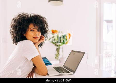 Porträt einer selbstbewussten jungen Frau mit lockigen Haaren mit Laptop auf dem Tisch zu Hause Stockfoto