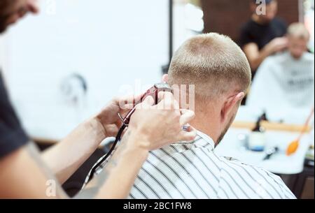 Die Rückansicht des Mannes im barber Shop hat seinen Nacken mit einem Trimmer rasiert, der Friseur hält ein rotes Werkzeug. Nahaufnahme Stockfoto