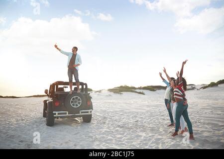 Junger Mann auf Jeep-Reifen, der selfie mit Freunden am Strand nimmt Stockfoto