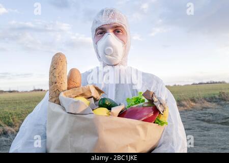 Porträt des Mannes mit Schutzanzug und Maske hält Lebensmitteltasche auf dem Land Stockfoto