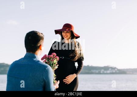 Junger Mann, der seiner schwangeren Freundin am Wasser Blumen schenkt Stockfoto