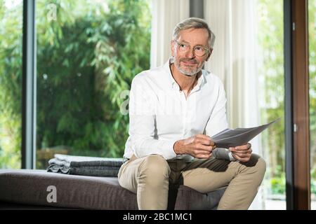 Lächelnder älterer Mann mit grauen Haaren im modernen Design Wohnzimmer, der auf der Couch sitzt und im Home Office auf Papier arbeitet Stockfoto