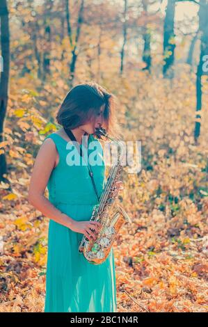 Süßes junges brünette Mädchen in einem langen blauen Kleid spielt im Herbstwald das gelbe Saxofon Stockfoto