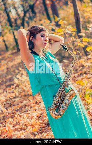 Süßes junges dunkelhaariges Mädchen mit Saxofon posiert in einem langen blauen Kleid in einem gelben Herbstwald und hebt die Hände zum Kopf Stockfoto