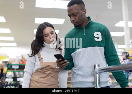 Weibliche Lebensmittelhändler helfen männlichen Kunden mit dem Smartphone im Supermarkt Stockfoto