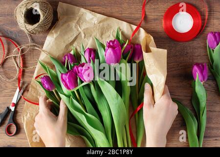 Frau Floristin wickelt Blumenstrauß von erstaunlichen lila Tulpen in Kunsthandwerk Papier auf Holztisch Draufsicht. Stockfoto