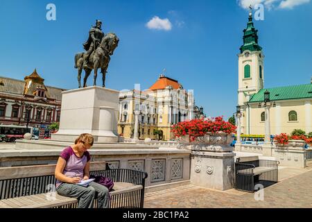 Eine Frau, die auf einer Bank sitzt und schreibt, umgeben von Architektur des Barock und der Sezessionskunst. Einheitsplatz in der Stadt Oradea, Rumänien. Juni 2017. Stockfoto