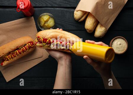 Person drückt Sauce auf heißen Hund auf Holzhintergrund, Draufsicht Stockfoto