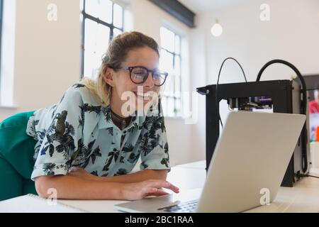Lächelnde Designerin, die am Laptop neben dem 3D-Drucker im Büro arbeitet Stockfoto