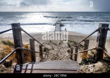 Verwitterte Holzstufen führen hinunter auf einen sandigen Strand, der zum breiten offenen Meer mit einem Holzsteg führt, der zu einer kleinen Felseninsel führt, einem Silhouette o Stockfoto