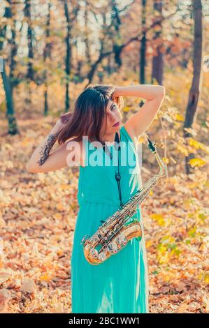 Sehr schöne junge Frau mit schwarzem Haar hob ihre Hände auf den Kopf und steht mit einem Altsaxophon in einem langen blauen Kleid unter den Bäumen in einem Ye Stockfoto