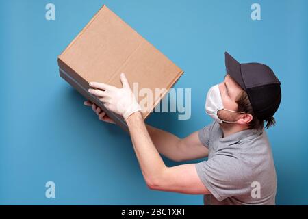 Kaukasischer junger Mann in medizinischer Maske, der ein Paket hält, das er dem Kunden gibt. Kein ansteckendes Lieferkonzept. Studio an blauer Wand gedreht. Stockfoto