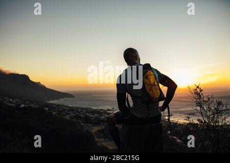 Rückansicht des athletischen Mannes, der auf einem Felsen sitzt und eine Pause nach dem laufen des Pfades bei Sonnenuntergang nimmt. Mann entspannt auf einem Felsen neben einem Pfad auf einem Moun sitzen Stockfoto