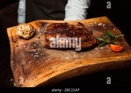 Auf einem Holzbrett liegt ein appetitliches, frisch zubereitetes gegrilltes Fleischsteak. Mit Salzstücken bestreut, daneben gibt es einen Rasmarin und eine Tomatenscheibe. Stockfoto