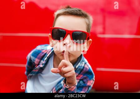 Junge in roter Sonnenbrille mit Finger auf den Lippen, was eine stille Geste in der Stadt macht Stockfoto