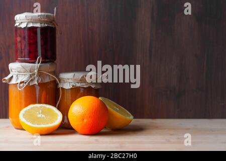 Gläser mit verschiedenen Marmeladen und Zitronenschnitt und Orange auf dunklem Holzhintergrund. Strowberry und Aprikosenmarmelade. Stockfoto