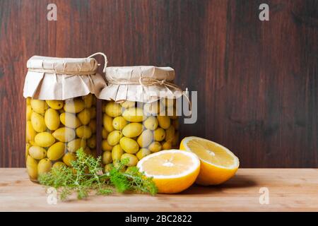 Konservierte fermentierte Oliven in Glasgefäßen mit wildem Fenchel und Zitronenschnitt auf dunklem Holzhintergrund. Gemüse Konserven. Stockfoto