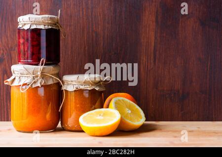 Gläser mit verschiedenen Marmeladen und Zitronenschnitt und Orange auf dunklem Holzhintergrund. Strowberry und Aprikosenmarmelade. Stockfoto