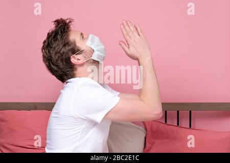 Kaukasischer Mann in medizinischer Maske, der die Hände zusammenhält und betet, während er allein im Bett in Quarantäne liegt. Studio gedreht. Stockfoto