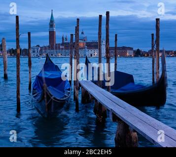 Blaue Stunde in Venedig: San Giorgio Maggiore Insel & Kirche über welligen Wasser, was dazu führt, dass sich Gondeln im Vordergrund bewegen und bei langer Belichtung verschwimmen Stockfoto