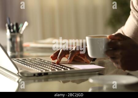 Nahaufnahme eines schwarzen Mannes mit einem Laptop, der nachts auf einem Schreibtisch zu Hause eine Tasse Kaffee hält Stockfoto