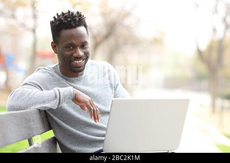 Fröhlicher schwarzer Mann mit Laptop und Blick auf die Kamera, die auf einer Parkbank sitzt Stockfoto