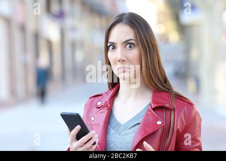 Ratlos beäugte Frau mit Handy schaut auf die Kamera, die auf der Straße steht Stockfoto