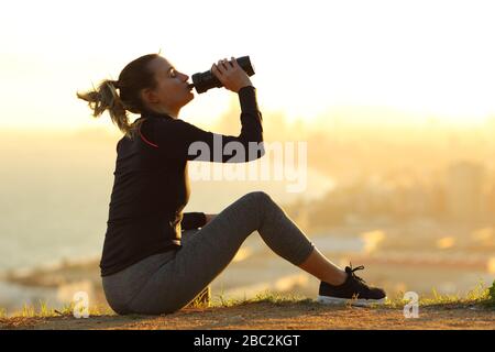 Seitenansicht Portrait eines ganzen Körpers einer Läuferin, die bei Sonnenuntergang am Stadtrand Trinkwasser aus einer Flasche stillt Stockfoto