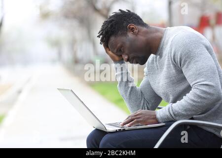 Trauriger schwarzer Mann, der sich beschwert, den Inhalt des Laptops auf einer Bank in einem Park zu überprüfen Stockfoto