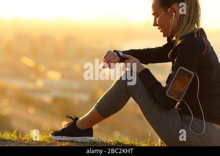 Der Läufer, der Kopfhörer trägt, überprüft die Smartwatch, die bei Sonnenuntergang auf dem Boden sitzt Stockfoto