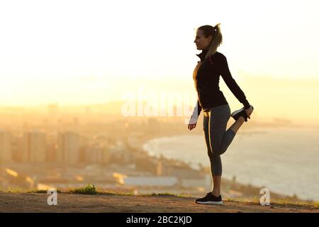 Seitenansicht Ganzkörperporträt einer Läuferin, die sich bei Sonnenuntergang am Stadtrand ein Bein streckt Stockfoto