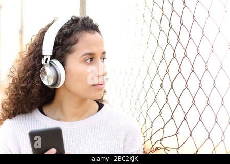 Das ernste Latina-Mädchen, das Musik hört und das Smartphone auf der Straße hält, blickt durch ein Gitter Stockfoto