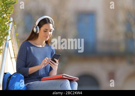 Studenten, die Kopfhörer tragen, hören Musik mit einem Smartphone, das im Freien auf einem Universitätsgelände sitzt Stockfoto