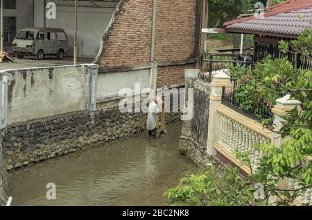 21. Juni 2018 Panyambungan, Sumatra, Indonesien: Lokaler armer Mann, der mit dem Netz in einem trügen schmutzigen Fluss mitten in der Stadt fischt, auch alter schmutziger Schabby-Wagen sichtbar Stockfoto