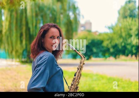 Rot-köpfiges junges Mädchen in blauer Jacke mit einem Saxofon lächelnd im Park Stockfoto