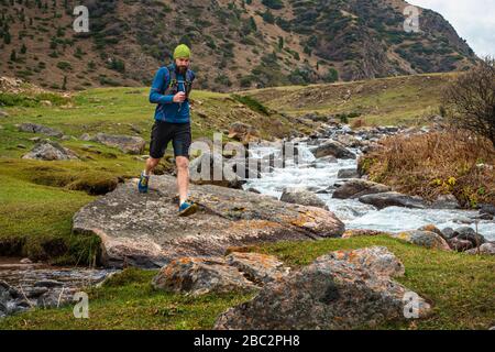 Runner Kerl joggt im Hochland. Athlet läuft in der Nähe eines Gebirgsflusses. Der Mann trainiert im Freien. Trail Running Stockfoto