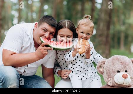 Urlaub im Freien Portrait von glücklichen Familien, die Wassermelone beim Picknick essen: Mutter, Vater und Tochter mit ihrem Teddy haben zusammen Spaß im Park Stockfoto