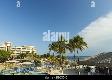 Schöner Blick auf die Hotelanlage. Außenpool mit blauen Sonnenliegen und Sonnenschirmen auf grünem Palmengrund. Willemstad. Curacao. Stockfoto