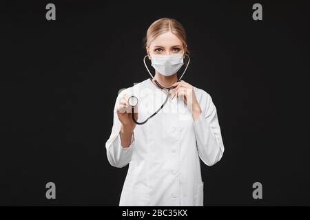 Junge Ärztin in medizinischer Uniform und Schutzmaske mit einem Stethoskop, isoliert am schwarzen Hintergrund mit Kopierraum. Gesundheitswesen und Stockfoto