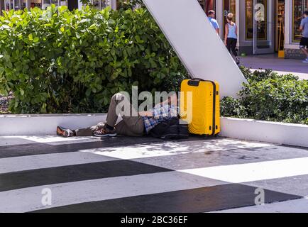 Blick auf den müden Mann mit gelbem Schlafsack auf dem Boden in der Nähe von grünen Büschen auf der Gehstraße. Miami. USA. Stockfoto
