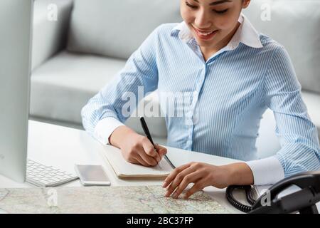 Der Reiseleiter sitzt am Tisch im Büro und schaut sich die Karte an, die Notizen gemacht hat. Er lächelt aus nächster Nähe Stockfoto