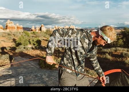 Mann mit Camo und Stirnband richtet Zelt bei Sonnenuntergang in der Wüste auf Stockfoto