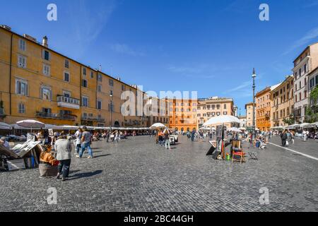 Ein sonniger Tag mit blauem Himmel auf der alten Piazza Navona, während Touristen ihre Kunstwerke und Gemälde besichtigen und an Straßenkünstlern vorbeiwandern. Stockfoto