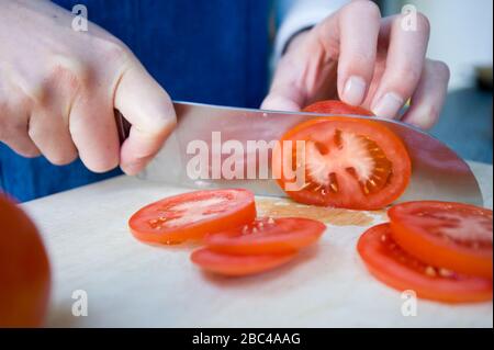 Ein Paar Hände, die ein scharfes Messer halten, um eine Tomate in Scheiben zu schneiden. Stockfoto