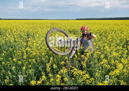 Radfahrerinnen bewegen sich zu Fuß durch ein Rapsfeld und heben ihr Mountainbike-Rad am Hinterrad an. Stockfoto